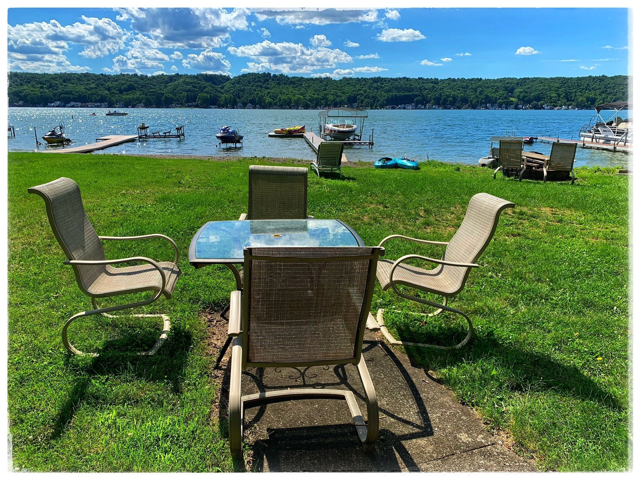 A grassy area with a glass table and four wicker chairs facing a lake with boats.
