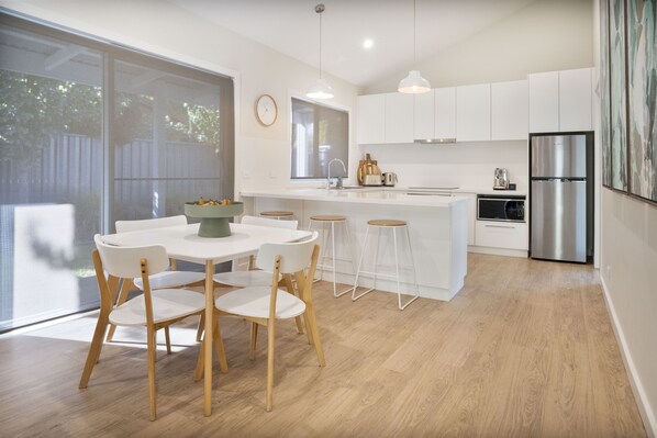 Una cocina moderna con armarios blancos, un refrigerador de acero inoxidable y un comedor con mesa y sillas de madera.