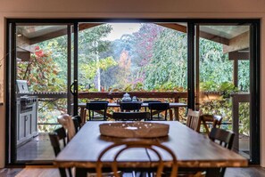 A dining area with a large table and chairs, a grill, and a view of a garden with trees and foliage. - Nest House - A picture speaks a thousand words.. (Bright)