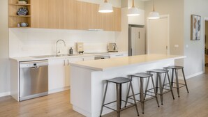 A modern kitchen with wooden cabinets, a stainless steel dishwasher, a large island with bar stools, pendant lighting, and a refrigerator.