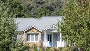 A two-story stone house with a blue door and white picket fence, surrounded by greenery and a hill in the background.