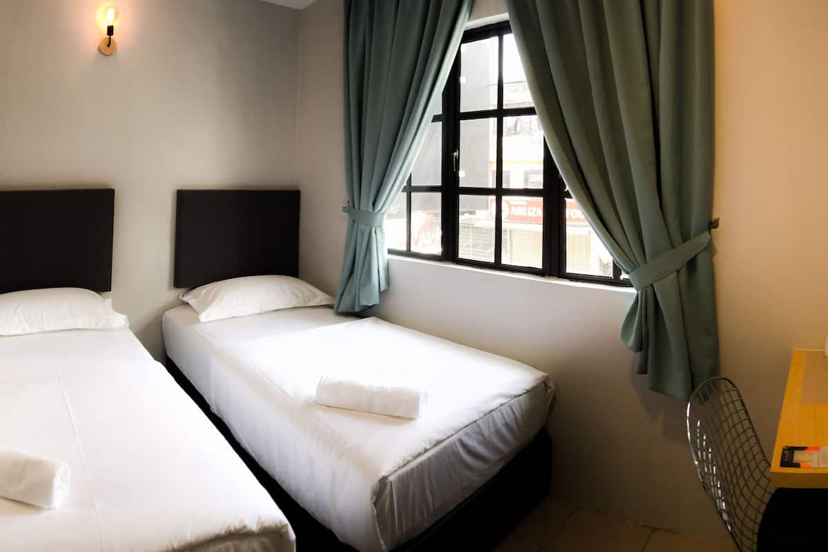 Ein Hotelzimmer mit zwei Einzelbetten, einem kleinen Tisch mit Wasserkocher und Fernbedienung sowie einem Fenster mit Vorhängen.