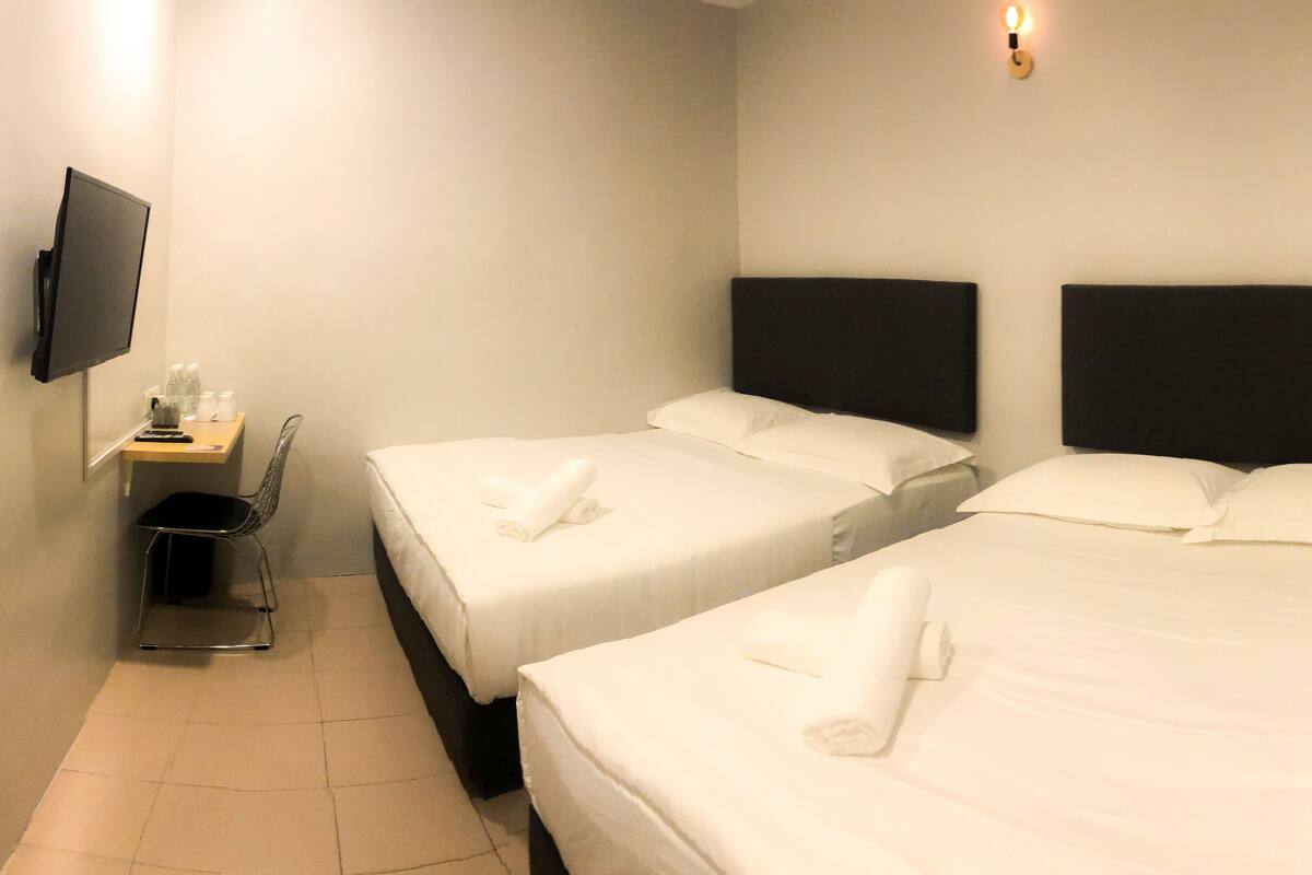Ein Hotelzimmer mit zwei Einzelbetten, einem kleinen Schreibtisch mit Stuhl, einem an der Wand befestigten Fernseher und einem Mülleimer.