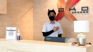 Un mostrador de recepción de hotel con una computadora portátil, un cartel con un código QR, un dispensador de desinfectante y una lámpara.