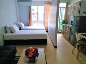 A bedroom with two beds, a small table with a flower vase, and a kitchenette with a refrigerator and cabinets. - DD Garden Apartment 2 (Bangkok)