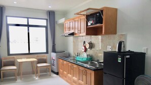 A compact kitchen with wooden cabinets, a black refrigerator, a microwave, and a fan. There is a window with curtains and a table with chairs. - Nhat Thu Hotel & Apartment (Ho Chi Minh City)