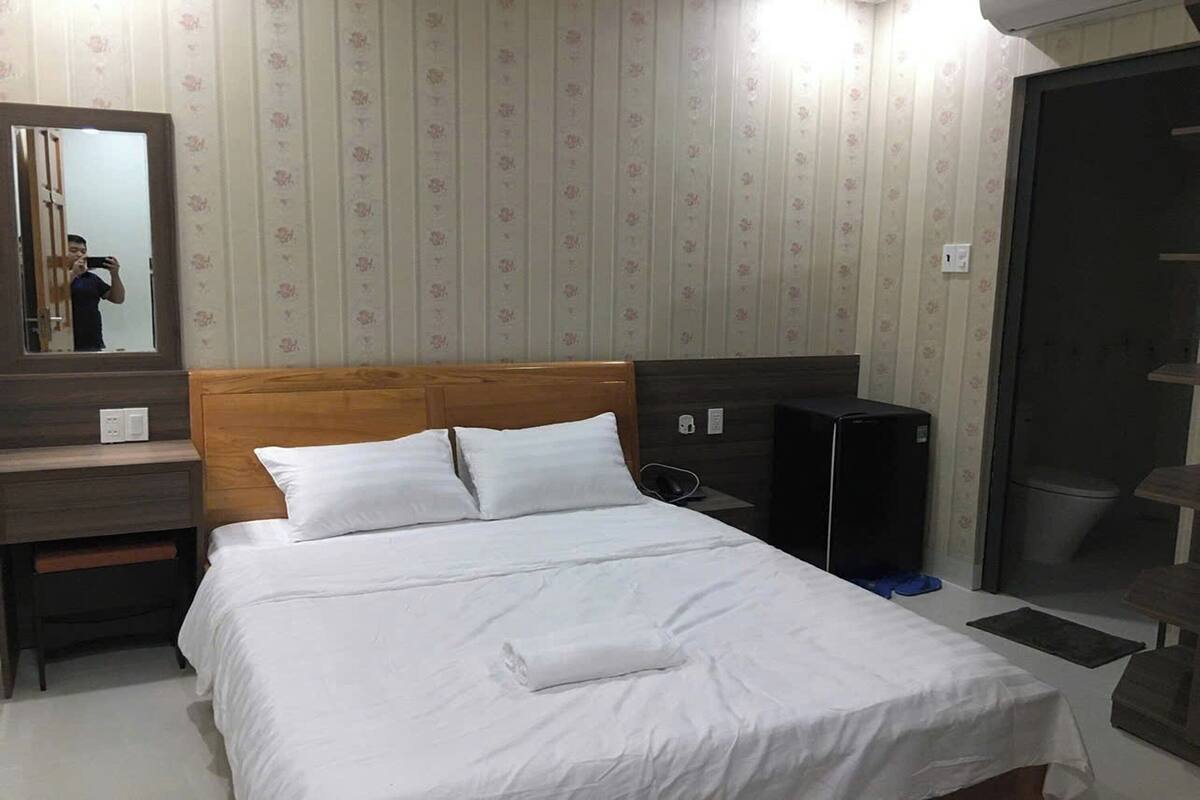 A hotel room with a wooden headboard, a large bed with white linens, a bedside table with a lamp, and a mirror.