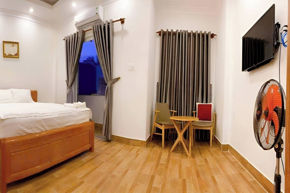 A bedroom with a wooden bed, a chair, a table, a TV, and a fan.