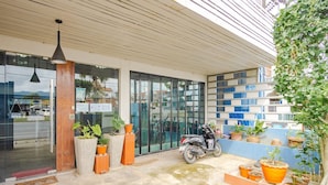 A modern building with a welcoming entrance, a scooter parked outside, and various potted plants arranged on the ground and along the walls. - Tyler Banpon (Si Sunthon)