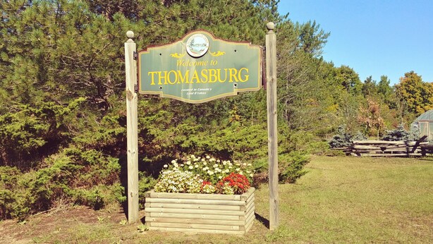 A welcome sign for Thomasburg, surrounded by a wooden planter with flowers, set against a backdrop of trees and a clear sky. - Escape to the Country! Sleeps 4 (Thomasburg)