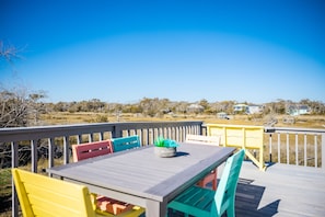 A wooden deck with a table and chairs, overlooking a grassy landscape with houses in the distance. - It Depends: Beach Cottage Overlooking Davis Canal (Oak Island)