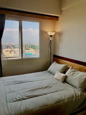 A hotel room with a bed, a window with a view of the city, and a floor lamp.