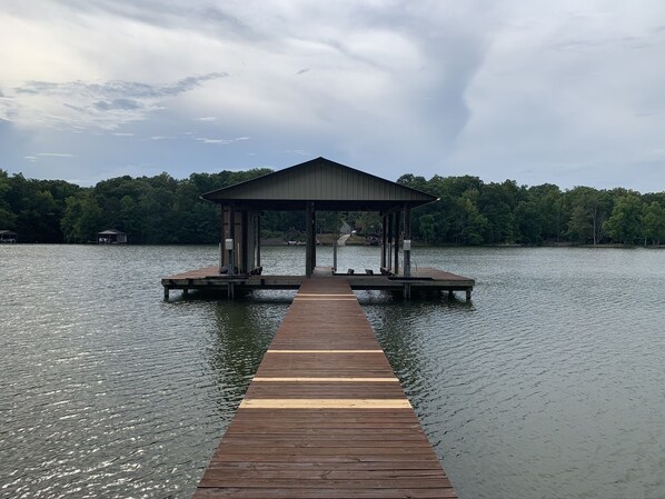 A wooden pier leading to a covered dock with a roof, surrounded by calm water and a forested shoreline.