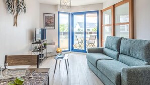 Un soggiorno moderno con un divano grigio, un tavolino rotondo e una TV su un mobile. È presente una grande finestra con cornici blu e vista sull'esterno.