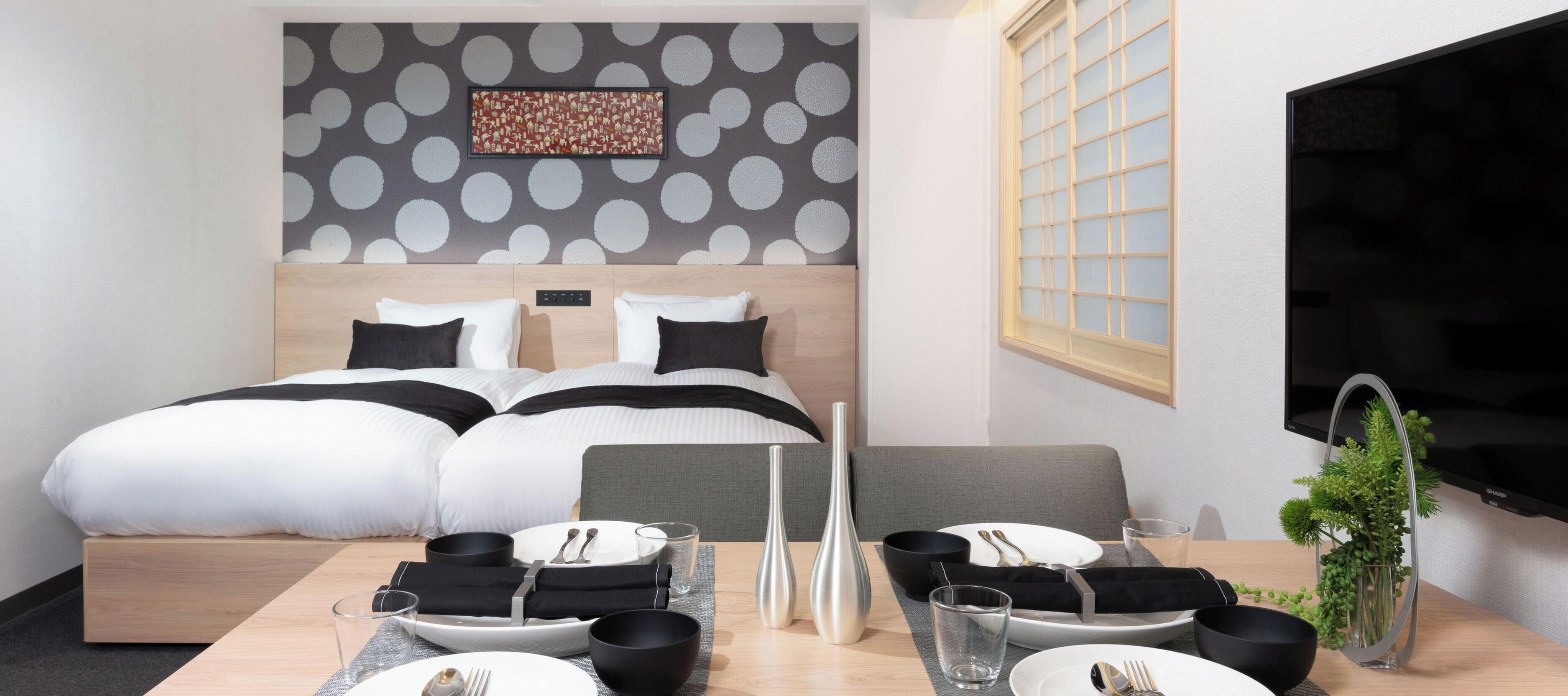 A hotel room with a bed, a dining table set with plates, bowls, and glasses, a TV, and a decorative wall piece.