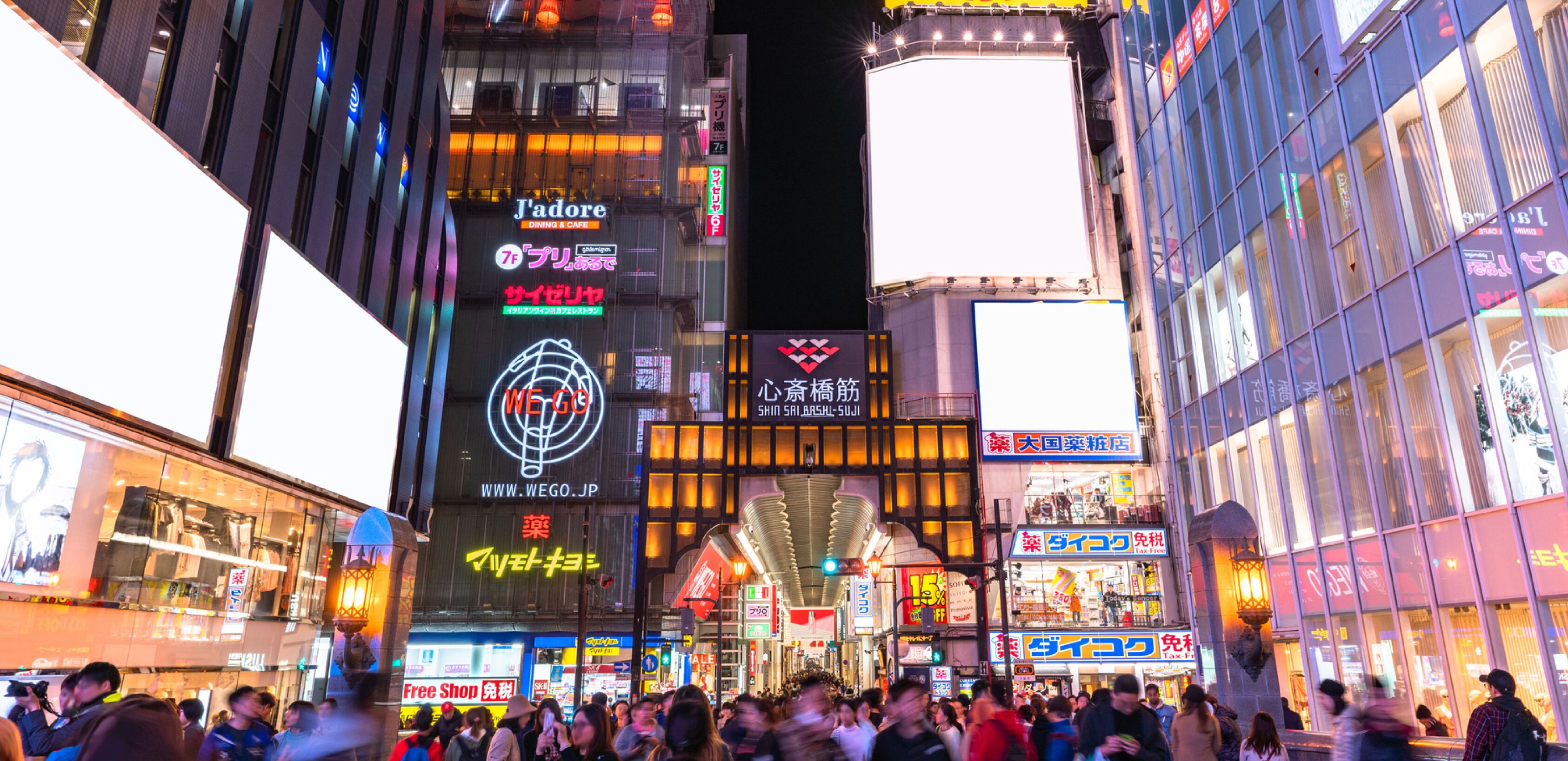 A busy urban street at night with illuminated signs and billboards, including a prominent 'WEGO' sign and a 'J'adoré' dining establishment.