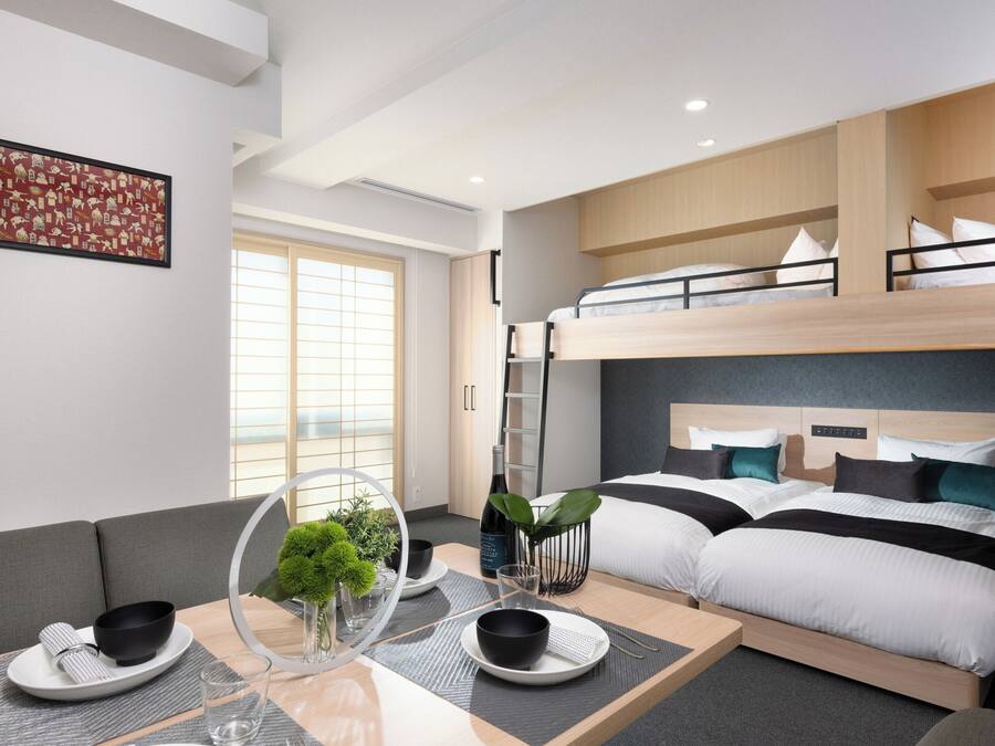 A modern hotel room with a bunk bed, a dining area with a table set for two, and a large window providing natural light.