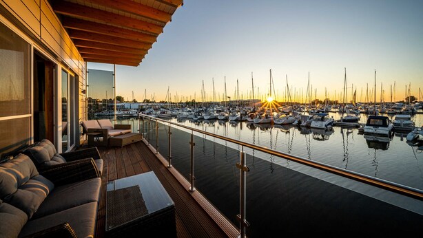 A marina with boats docked, a wooden deck with seating, and a sunset view. - 11. floating houses (105 m²) Annea with fireplace - Floating House Annea with fireplace (Kröslin)