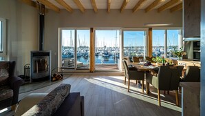 A spacious living area with a large window overlooking a marina, a fireplace, and a dining area with natural light. - 11. floating houses (105 m²) Annea with fireplace - Floating House Annea with fireplace (Kröslin)