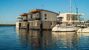 A modern houseboat with a balcony, large windows, and a sailboat docked nearby. - 11. floating houses (105 m²) Annea with fireplace - Floating House Annea with fireplace (Kröslin)