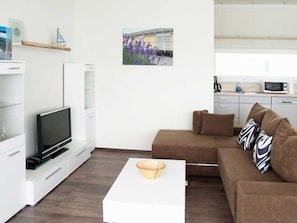 Una sala moderna con un televisor de pantalla plana, una mesa de centro blanca y un sofá marrón con cojines estampados. Encima del televisor, hay una estantería pequeña con objetos decorativos.