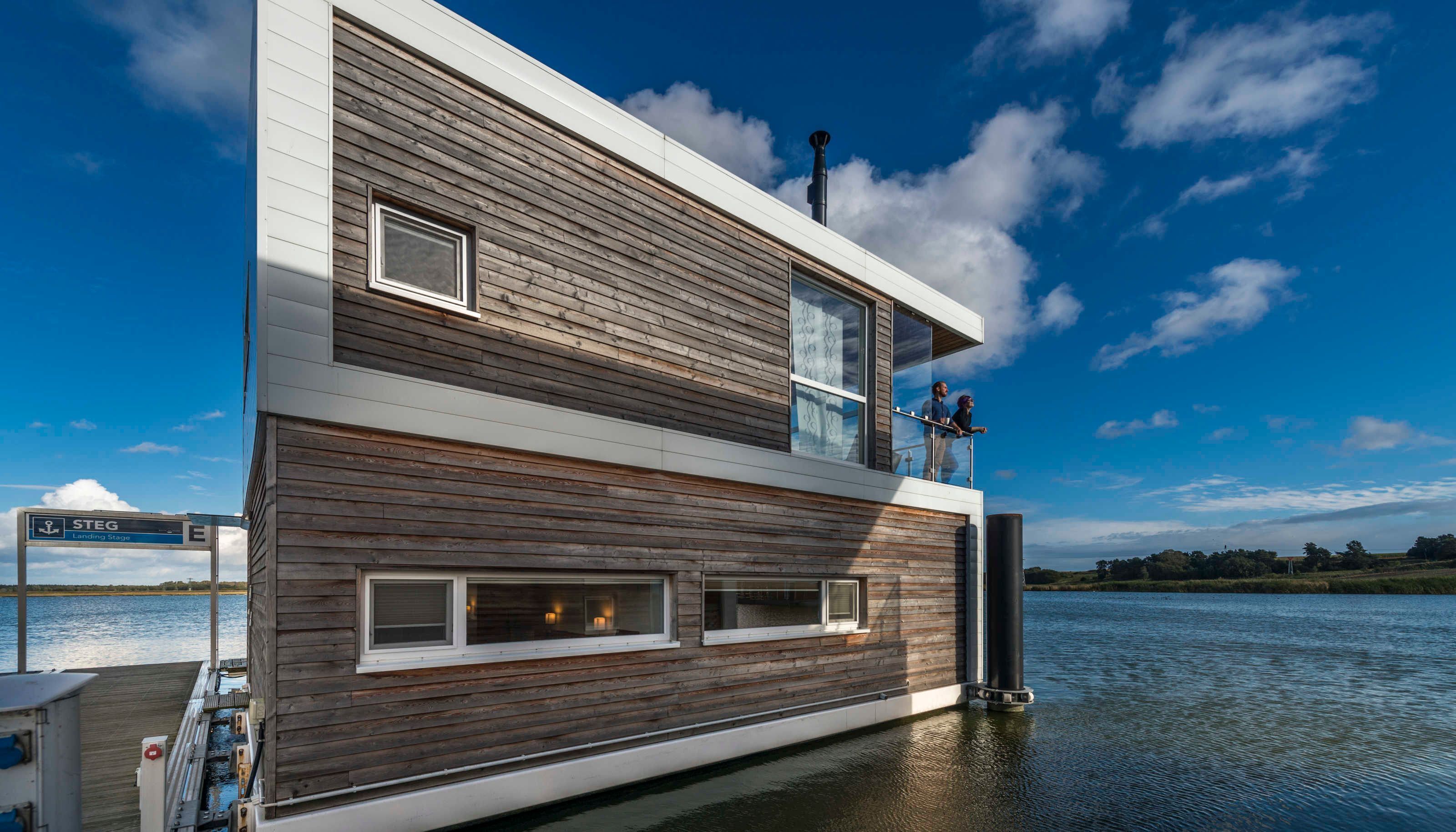 A modern houseboat with a wooden exterior, a balcony with two people, and a sign indicating a landing stage.
