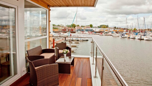 Une terrasse en bois, meublée de mobilier en osier, donnant sur une marina où sont amarrés des bateaux.