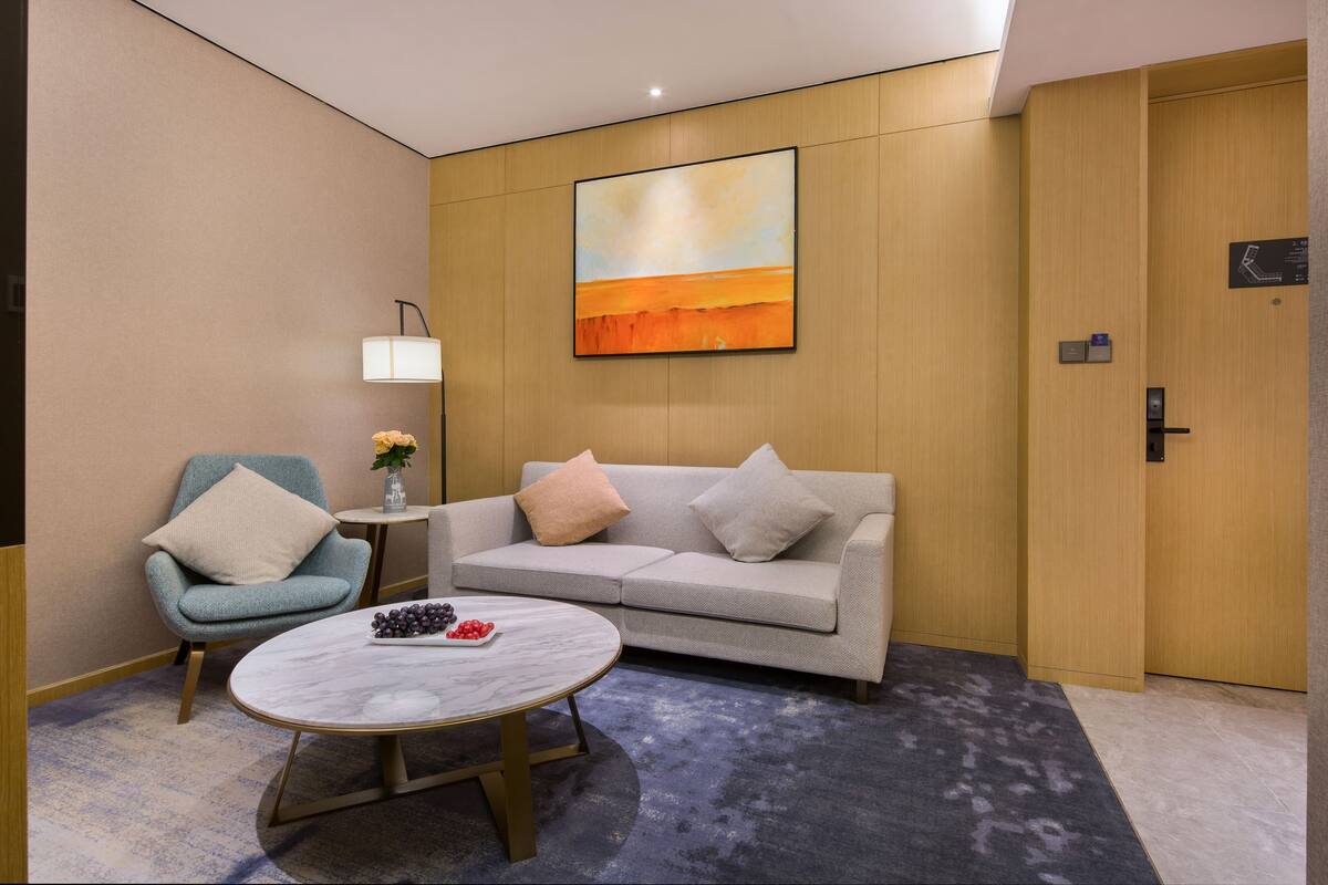 Una habitación de hotel moderna con sofá, sillón y una mesa redonda con fruta.