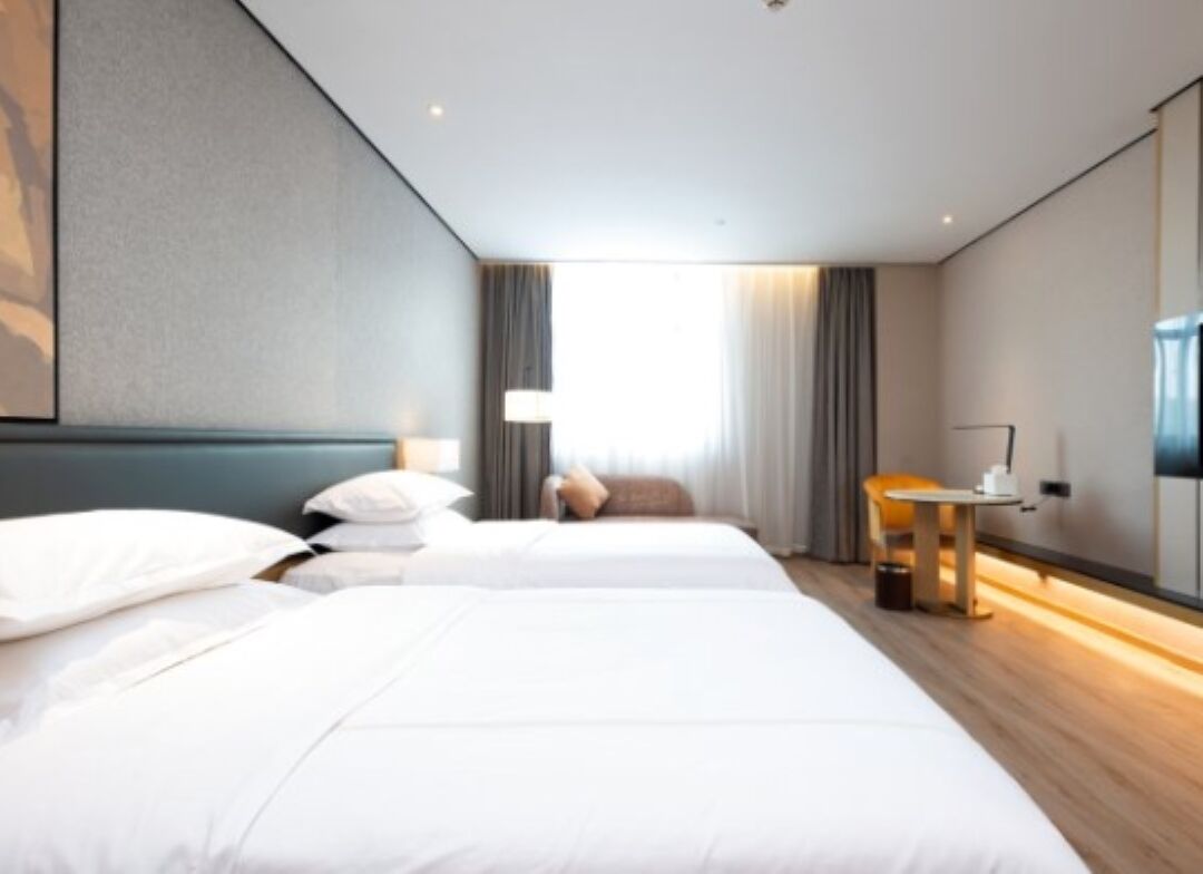 A hotel room with two beds, a desk, a chair, a lamp, and a large window with curtains.