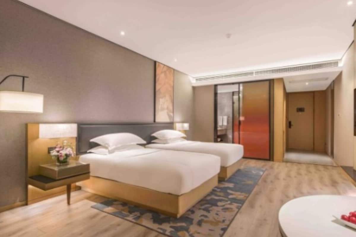 Una habitación de hotel moderna con dos camas, una mesita de noche con lámpara, un espejo grande y una mesa redonda con un jarrón de flores.