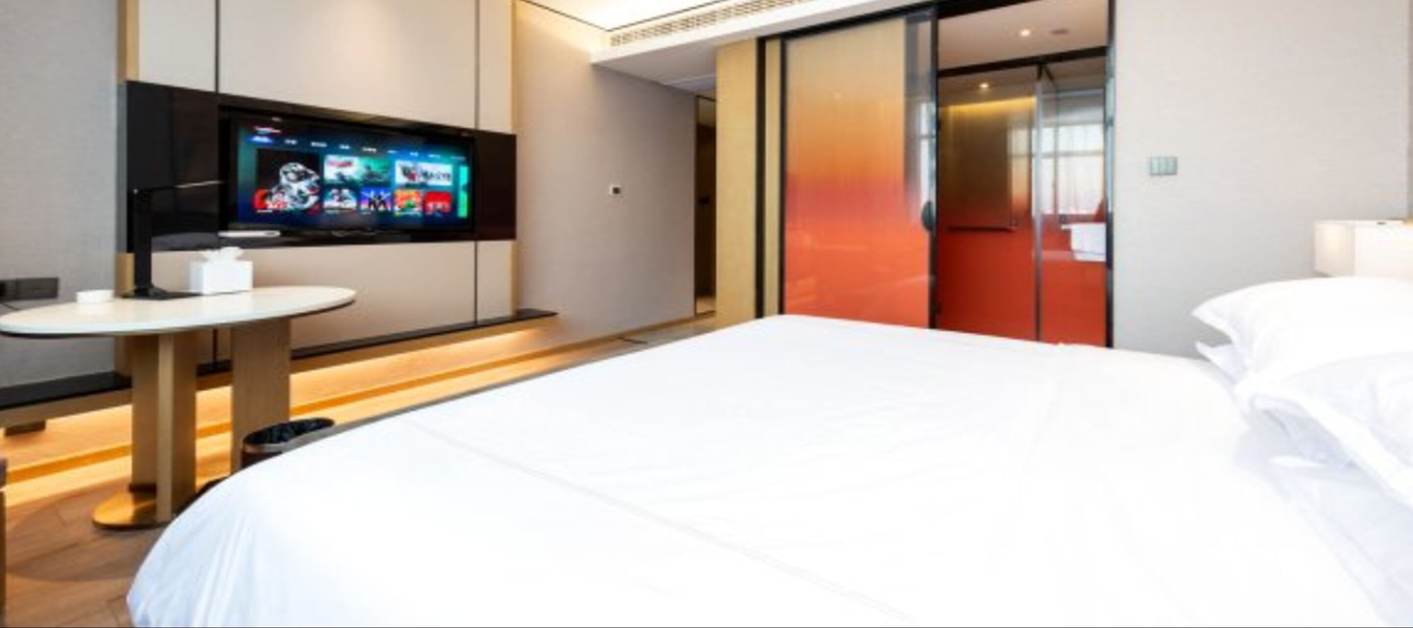 A hotel room with a large bed, a flat-screen TV mounted on the wall, a small round table, and a red accent wall.