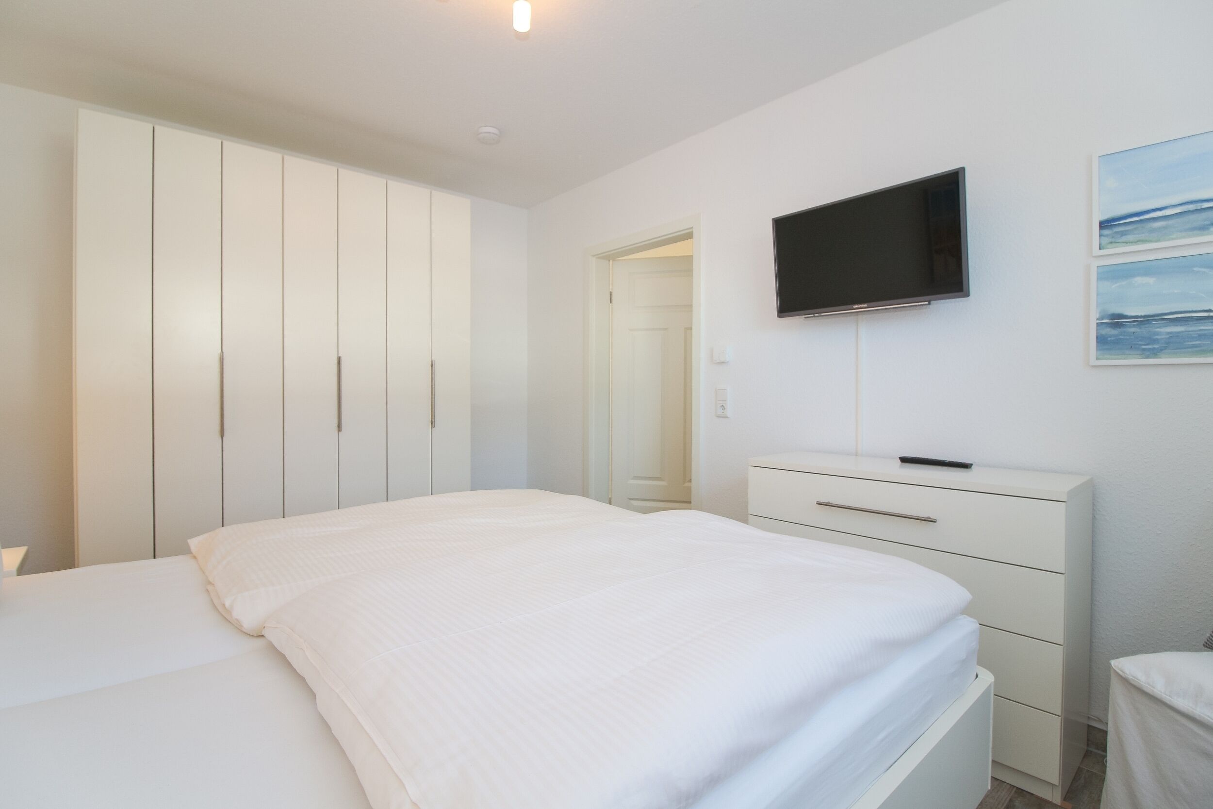 A bedroom with a bed, a dresser, a TV mounted on the wall, and a door leading to another room.