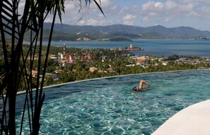 A swimming pool with clear water, a person swimming, and a view of a coastal town with buildings and mountains in the background.