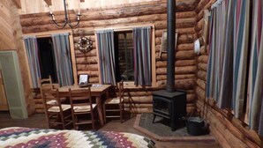 L’intérieur d’une cabane en rondins, avec une table à manger en bois, des chaises, un lit recouvert d’une courtepointe, une cheminée et des rideaux.