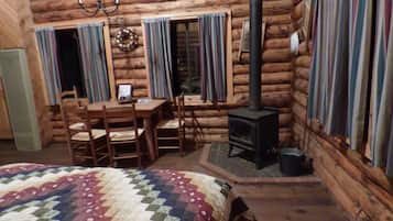 Interno di una baita di legno con un tavolo da pranzo in legno, sedie, un letto trapuntato, un camino e tende.