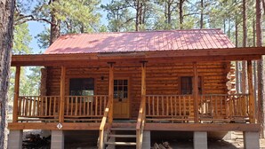 Une cabane en rondins surmontée d’un toit en métal rouge, une véranda à l’avant avec une balustrade en bois et un escalier menant à l’entrée.