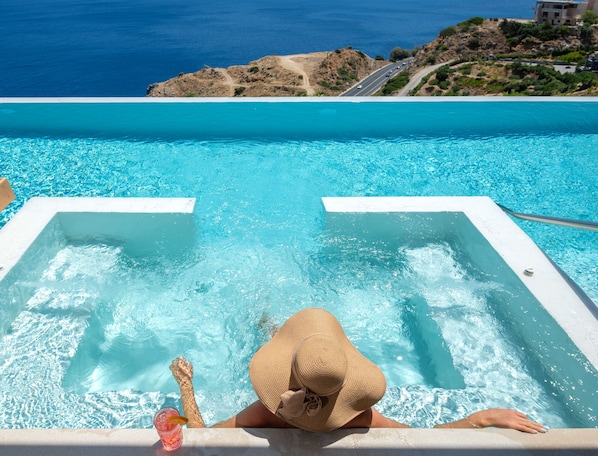 A pool with clear blue water, a person relaxing in a hot tub, and a view of the sea and a road in the background.