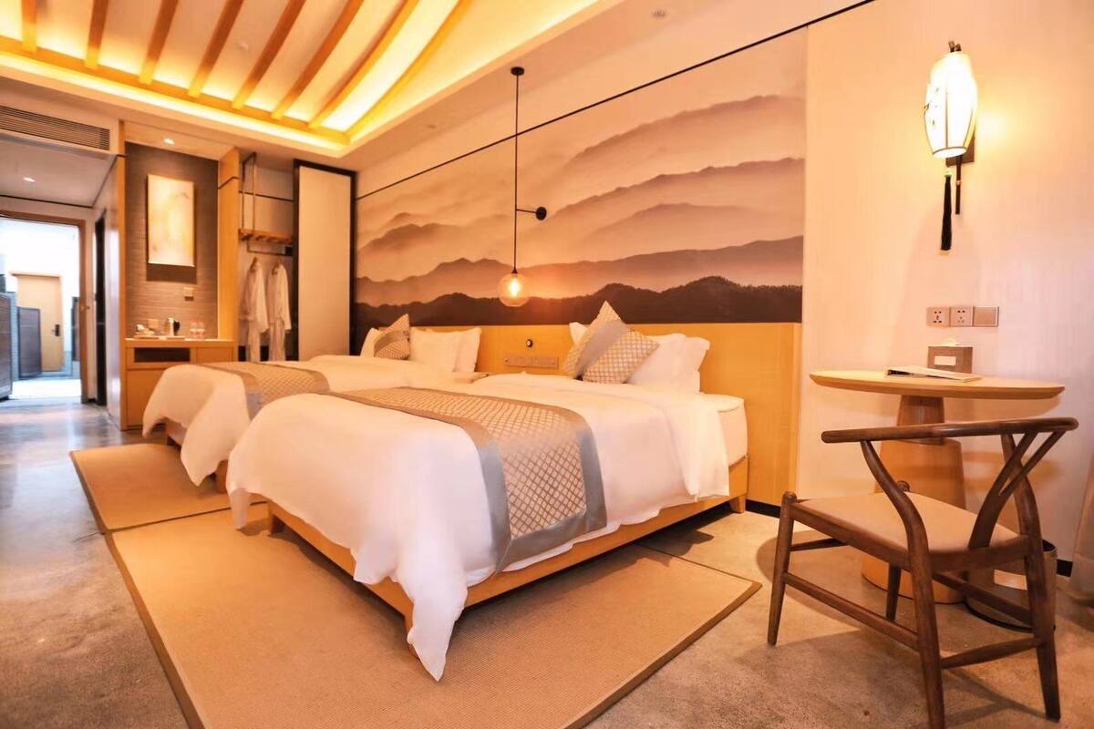 Habitación de hotel con una cama grande, una mesita y una silla. Se aprecia una pared con una pintura de montañas y una lámpara.