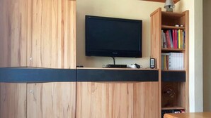 A modern room with a flat-screen TV mounted on a wooden cabinet, a bookshelf with books, and a small table with electronic devices. - Ferienhaus Ainsen (Bad Fuessing)
