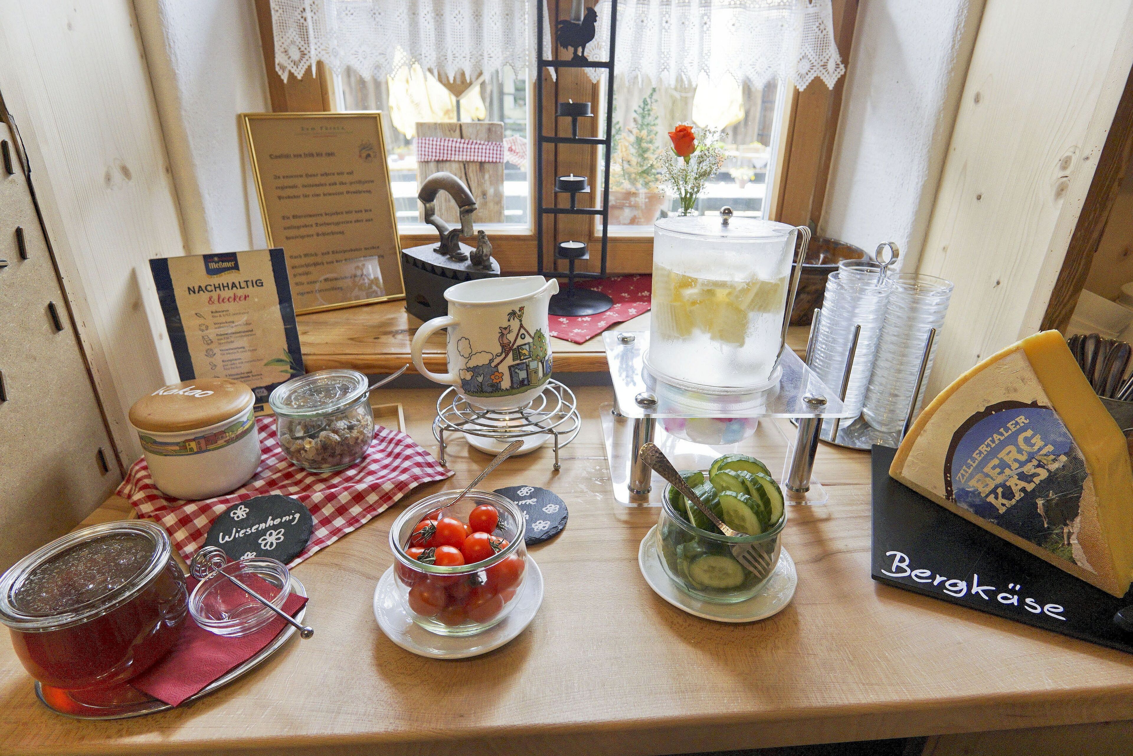 Una mesa de desayuno con diversos alimentos y condimentos, incluyendo una tabla de quesos, una jarrón y frascos de conservas.