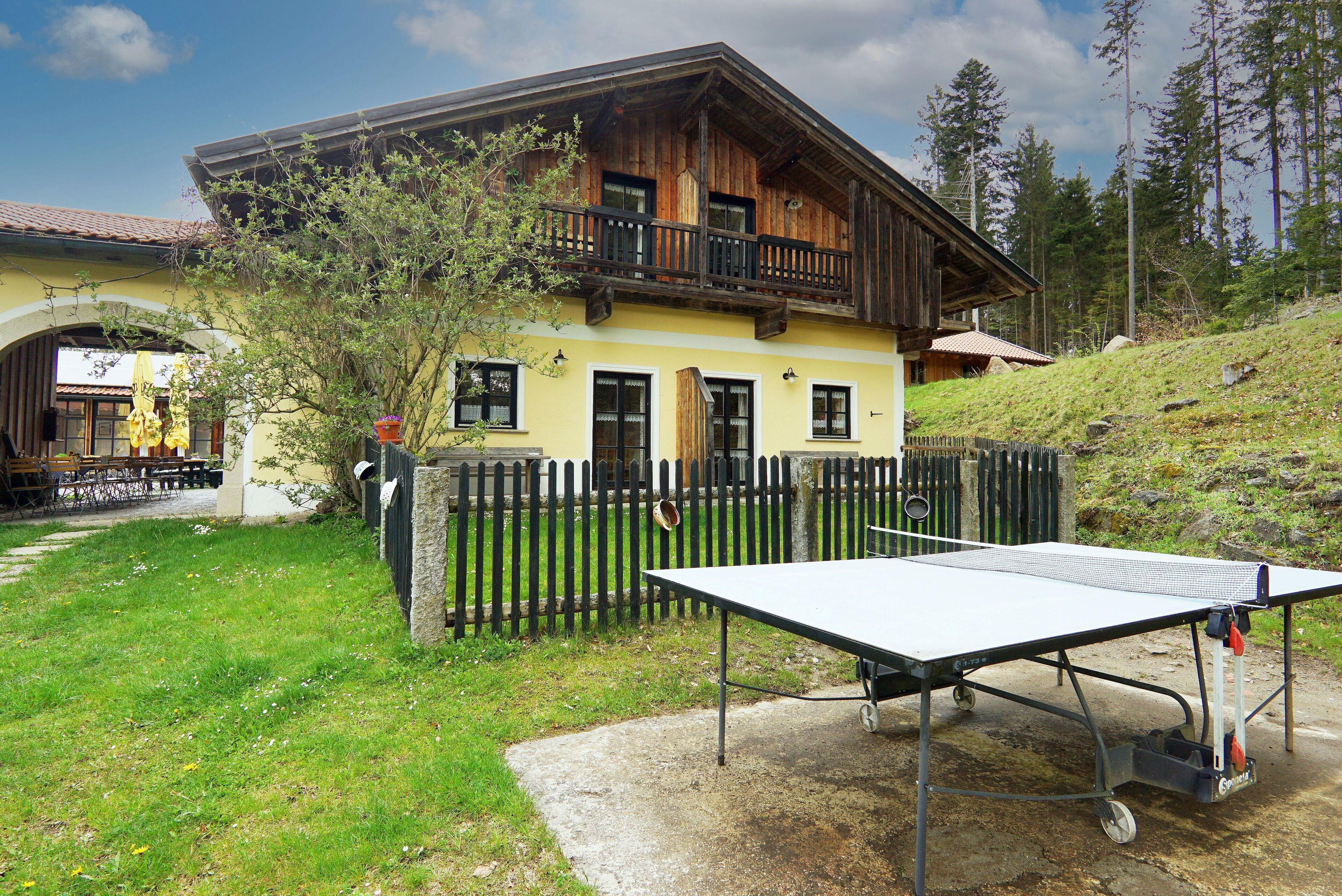 A wooden house with a balcony, a tennis table, and a fenced yard.