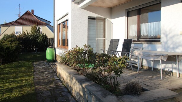 A residential building with a patio area featuring a table and chairs, a green barrel, and a stone pathway. - Ferienwohnung Friedrich (Schonungen)