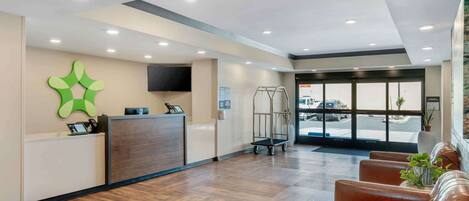 A reception area with a wooden floor, a reception desk, a green wall decoration, a television, and a view of the parking lot through the glass doors.
