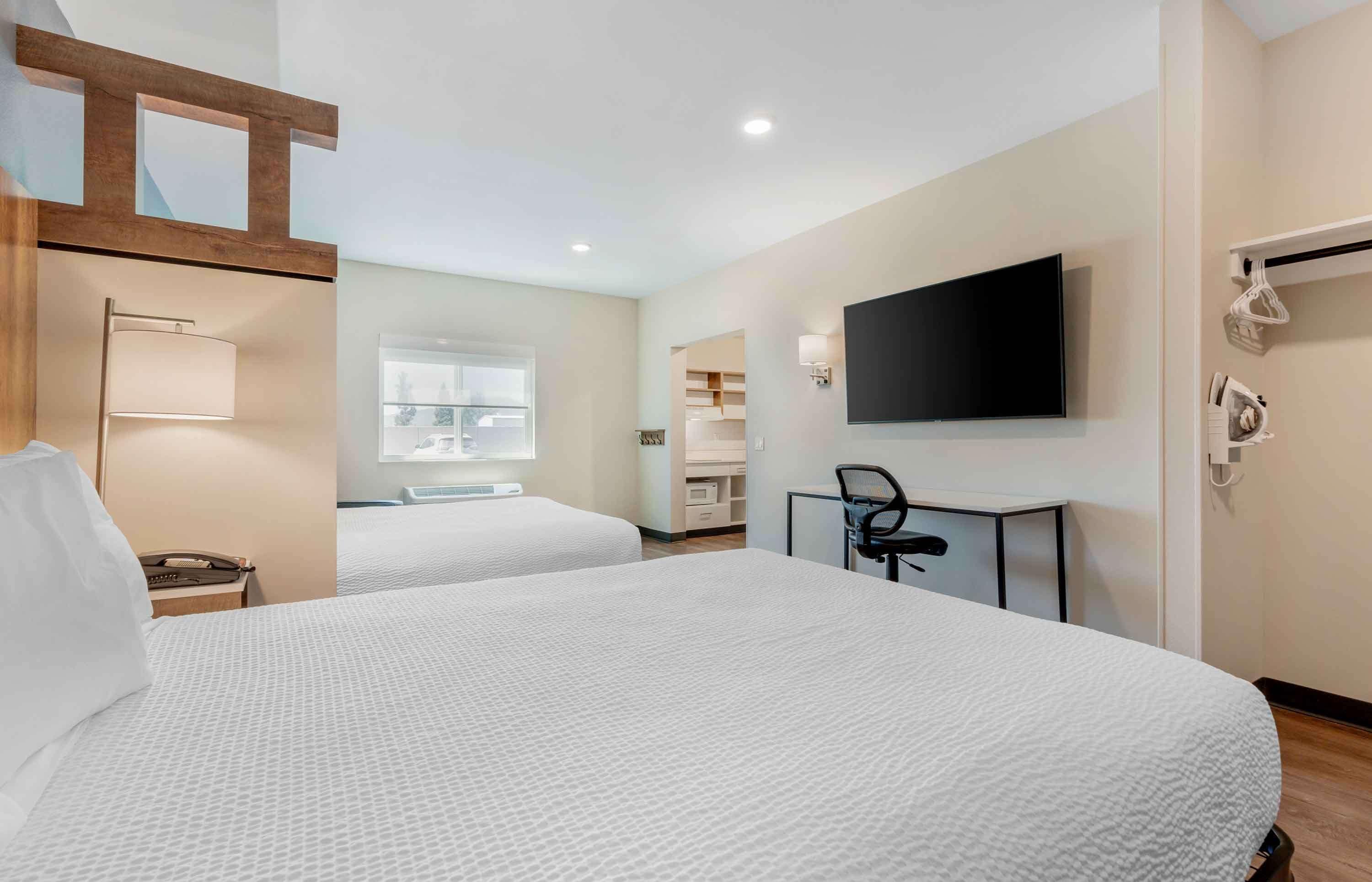 A hotel room with two beds, a TV mounted on the wall, a desk with a chair, and a window providing natural light.