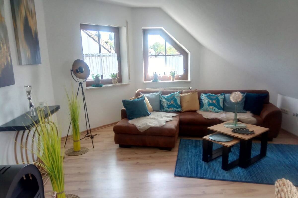 A living room with a brown sofa, blue and white cushions, a small wooden coffee table, and a blue rug. There are two windows with curtains, a floor lamp, and a plant.