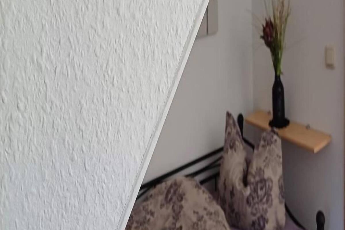 A bed with a patterned bedspread and pillows, a wooden shelf with a vase, and a white textured wall.