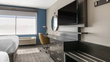 A modern hotel room with a bed, a desk, a chair, a flat-screen TV mounted on the wall, and a large window providing a view of the outside.