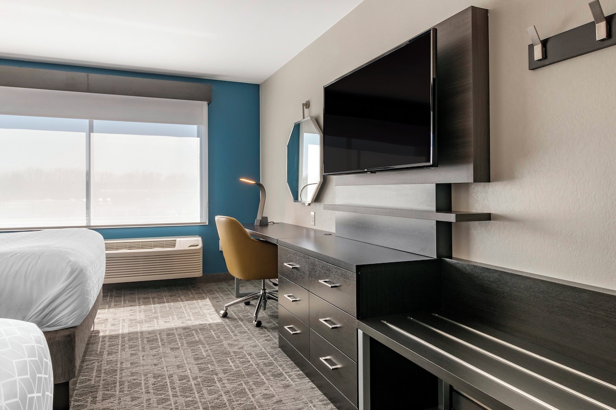 A modern hotel room with a bed, a desk with drawers, a chair, a flat-screen TV mounted on the wall, and a large window.
