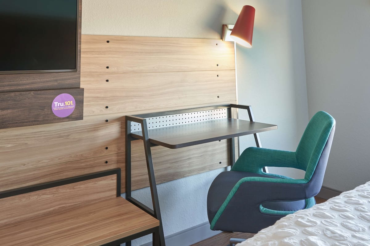 A hotel room with a wooden headboard, a desk with a pegboard, a green and gray chair, and a red lamp.