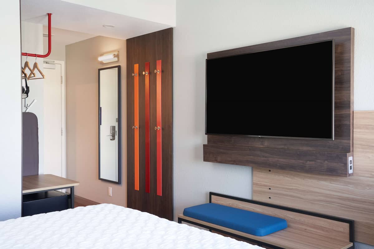 A modern hotel room with a bed, a desk, a TV mounted on the wall, and a wardrobe with sliding doors.